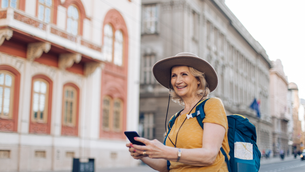 A smiling older woman with a backpack and hat looking at her phone on a European city street, representing walking tours for women over 50.